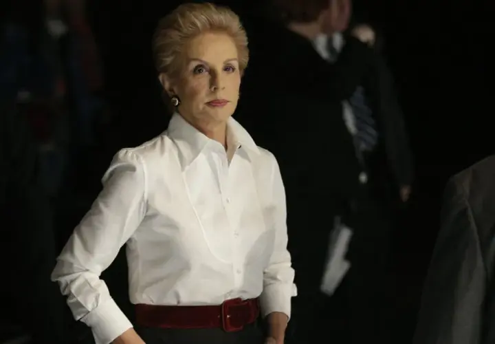 Elegancka Carolina Herrera w białej koszuli i czerwonym pasie, otoczona przez ludzi w ciemnych strojach.