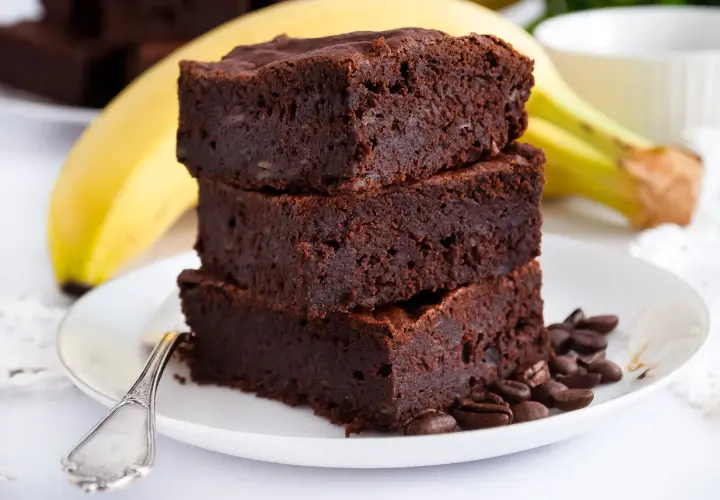 Stos puszystych brownie bananowych na talerzyku, obok dojrzały banan i ziarna kawy.