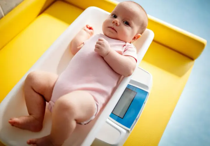 Noworodek w różowym body leży na wadze. Rodzice często zastanawiają się, ile powinien przybierać noworodek, by mieć pewność, że rozwija się prawidłowo.