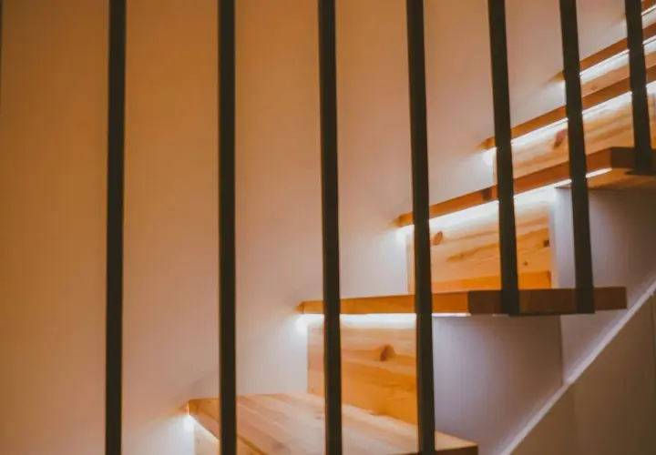 Drewniane stopnie schodów z subtelnym oświetleniem LED w ścianie. Czarne, pionowe balustrady tworzą nowoczesny kontrast.