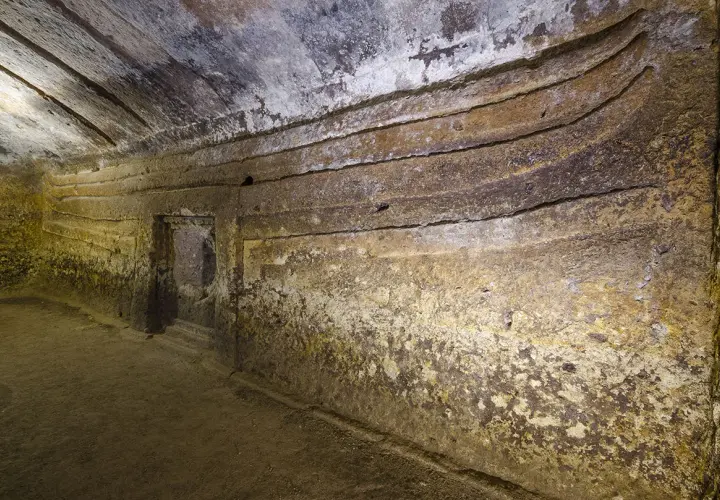 Interno di una domus de janas s'incantu, con pareti scavate e gradini che conducono a un'apertura.
