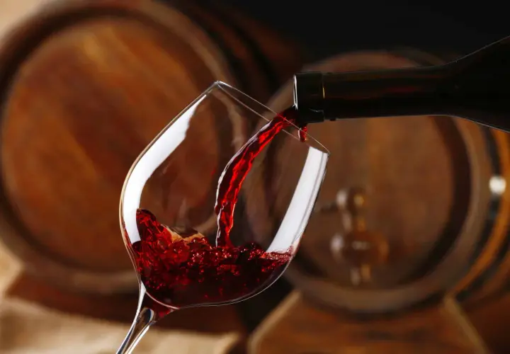 Czerwone wino, bogate w taniny, leje się z butelki do kieliszka, na tle drewnianych beczek.