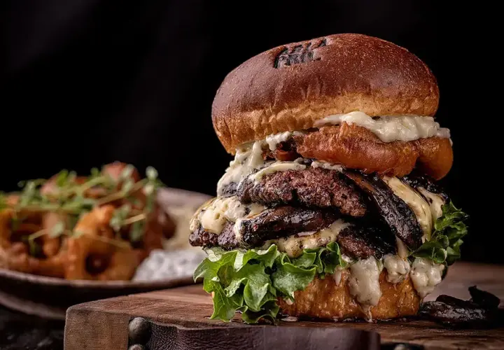 Un généreux burger maison avec plusieurs steaks, fromage fondu et laitue fraîche, servi sur une planche en bois.