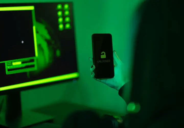 Ręka trzyma odblokowany telefon na tle monitora z zielonym kodem. Jak zrobić, by policja nie namierzyła telefonu?
