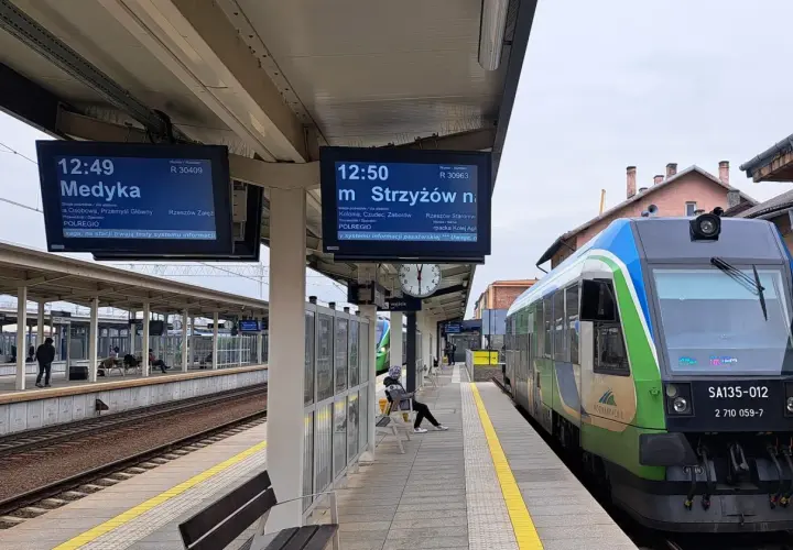 Na peronie kolejowym widać tablice informacyjne z rozkładem jazdy, w tym wskaźnik w4 pokazujący odjazd pociągu do Medyki. Obok stoi zielono-niebieski pociąg.