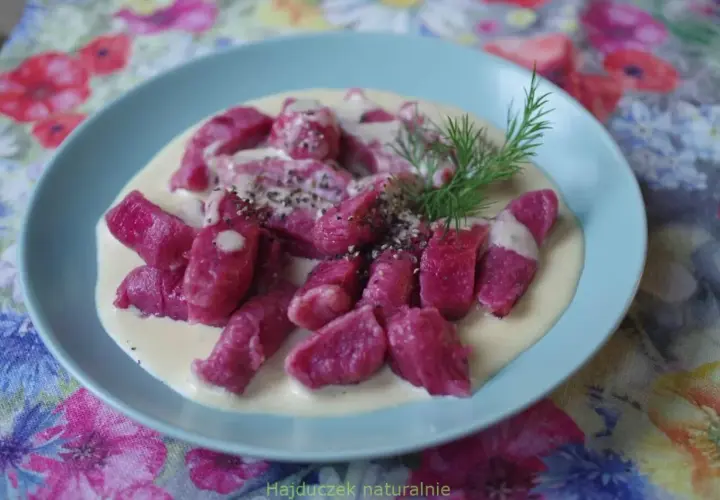 Różowe gnocchi buraczane w kremowym sosie, posypane pieprzem i udekorowane gałązką koperku, na talerzu z kwiecistym obrusem.