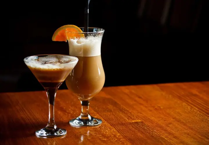 Dwa drinki na drewnianym stole: w kieliszku martini kawa z alkoholem i w wysokiej szklance latte z plasterkiem pomarańczy.