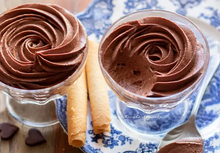 Due coppette di mousse al cioccolato ricetta classica, guarnite con rotoli di wafer e cuoricini di cioccolato.
