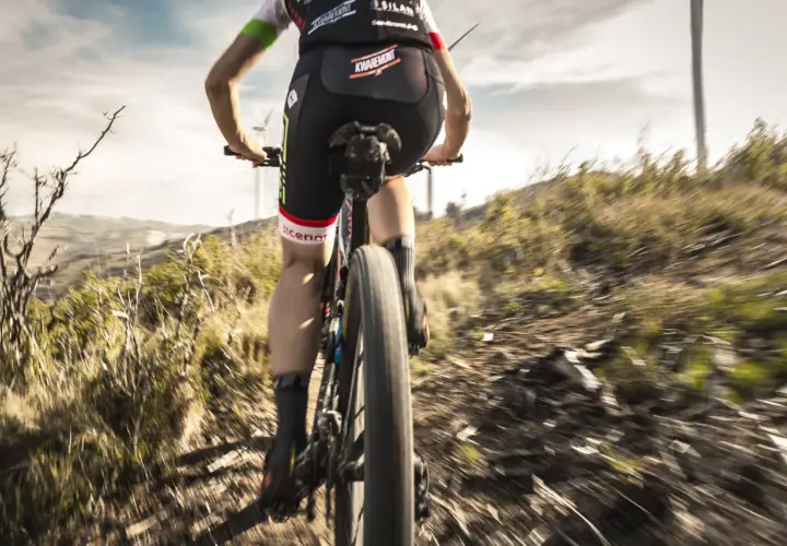 Ciclista desciende por un sendero polvoriento, disfrutando de una de las mejores rutas del mundo en bicicleta. El paisaje árido y las turbinas eólicas al fondo crean una escena épica.