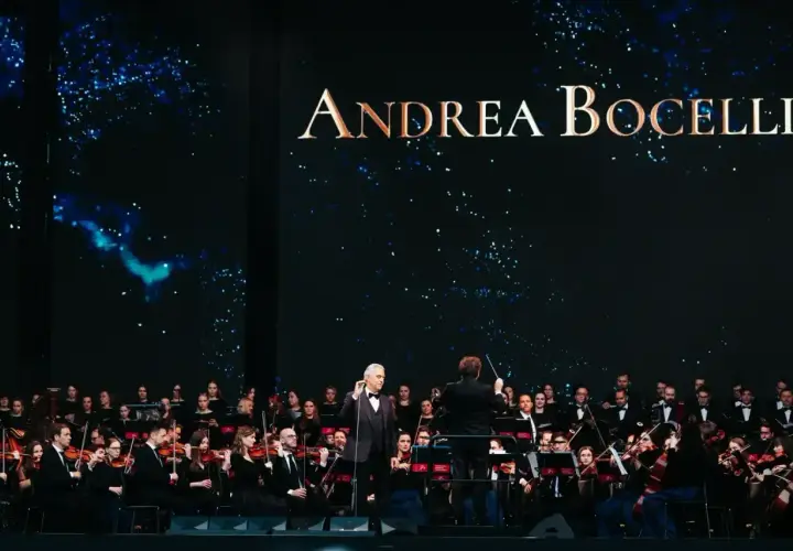 Andrea Bocelli koncert z orkiestrą symfoniczną. Wokalista w garniturze stoi na scenie, dyrygent kieruje muzykami.