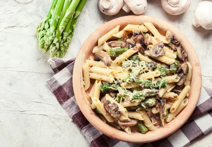 Penne z kremowym sosem, szparagami i pieczarkami, posypane parmezanem. Obok pęczek świeżych szparagów i kilka pieczarek.