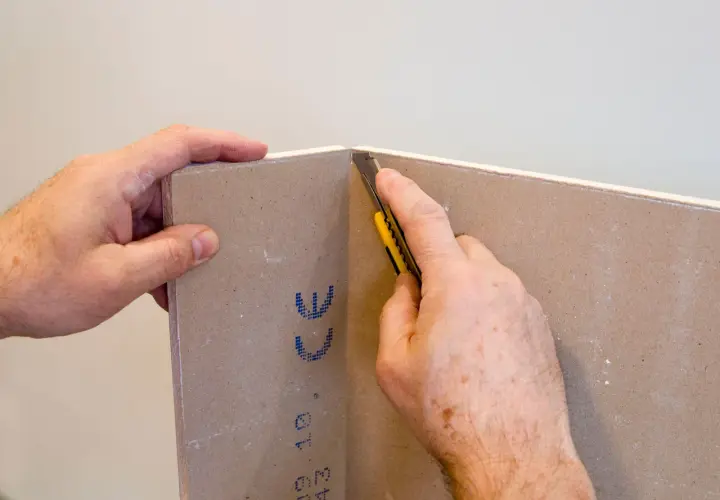 Mani che tengono un cartongesso mentre un taglierino viene usato per tagliare il materiale. Impara come tagliare il cartongesso con precisione.
