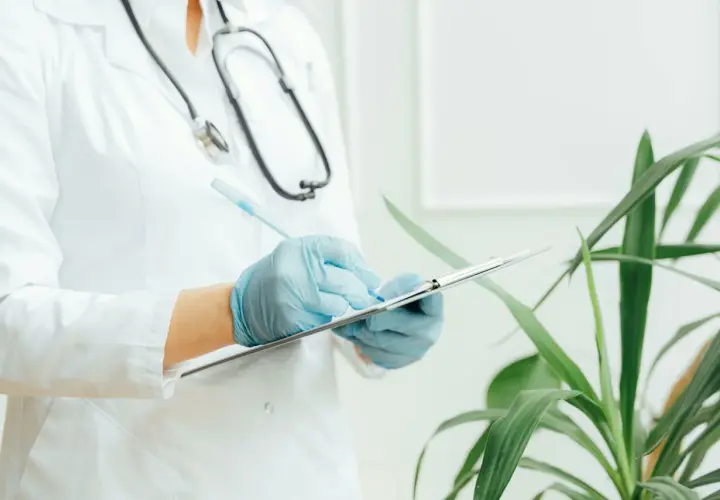 Medico con camice bianco e guanti blu scrive su un blocco appunti. Lo stetoscopio è appeso al collo. Il significato di "extramoenia" è legato a ciò che è fuori dalle mura, come un consulto medico fuori dall'ospedale.