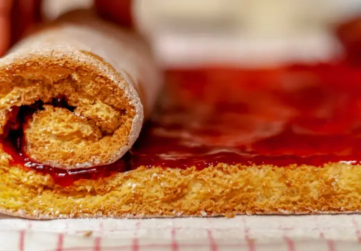Un rouleau de génoise moelleuse, saupoudré de sucre glace, est en train de rouler sur une couche de confiture de fraises éclatante.