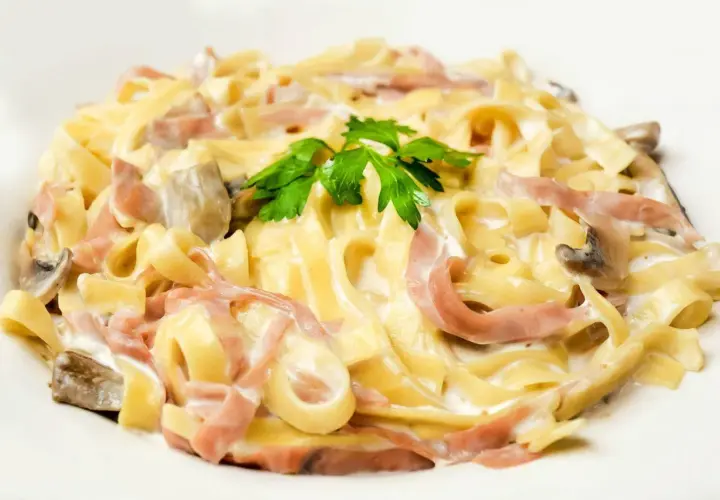 Tagliatelle z szynką i pieczarkami w kremowym sosie, z dodatkiem mozzarelli. Udekorowane natką pietruszki.