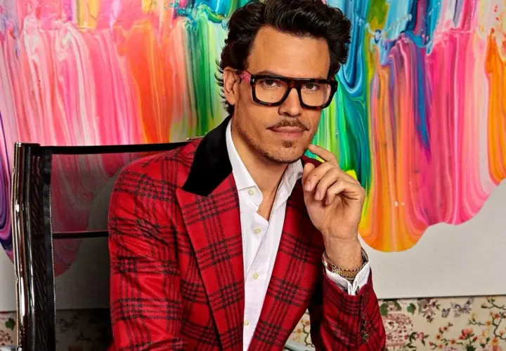 Juan Avellaneda, con su característico traje a cuadros rojos y gafas llamativas, posa con una mano en la barbilla frente a un vibrante cuadro abstracto.