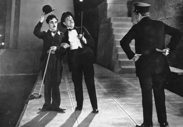 Charlie Chaplin i jego towarzysz w eleganckich strojach, z kapeluszami i laskami, w scenie z okresu kina niemego. Policjant stoi tyłem.