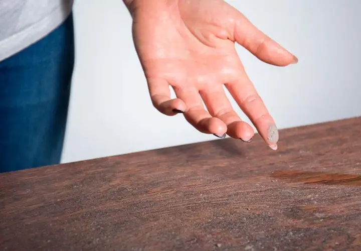 Mano con polvo en los dedos sobre una mesa de madera, indicando la necesidad de limpiar el polvo.