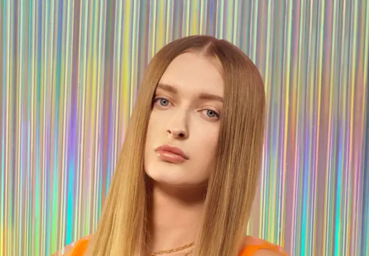 Sophie, top model o długich, blond włosach, pozuje na tle błyszczącej, tęczowej kurtyny.