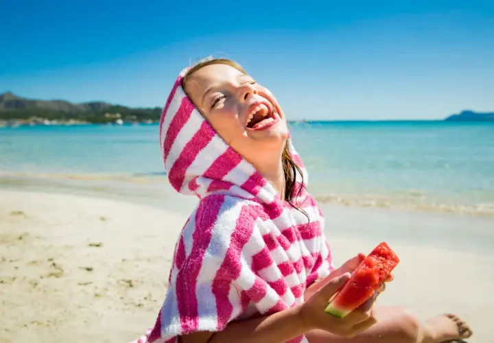 Niña riendo en la playa con una toalla a rayas y sandía, perfecta comida para llevar a la playa con niños.