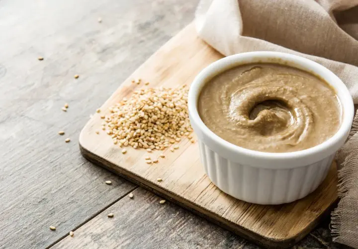 Domowe tahini jak zrobić? Ziarna sezamu i gotowa pasta na desce.