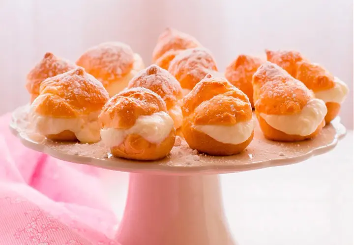 Deliciosos profiteroles de nata, espolvoreados con azúcar glas, listos para ser disfrutados en una bandeja blanca.