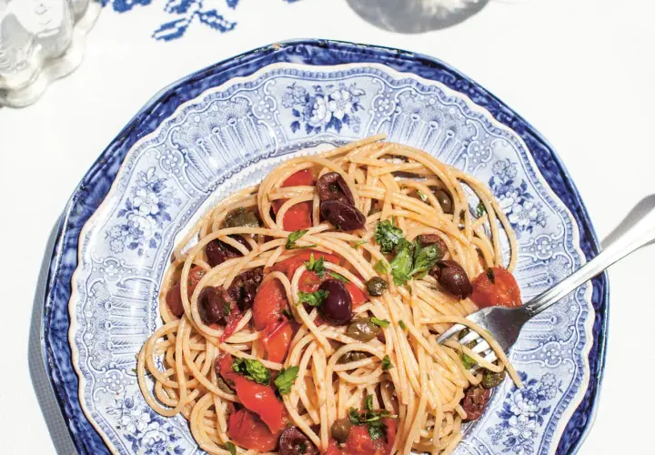 Spaghetti Puttanesca mit Oliven, Kapern und Tomaten auf einem blau-weißen Teller.
