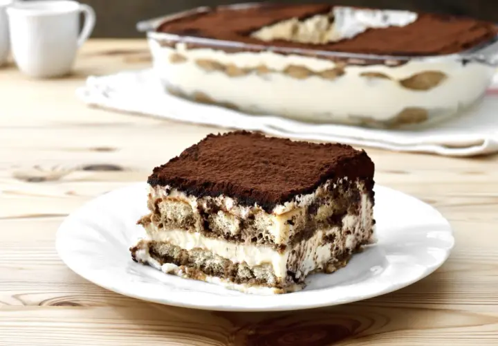 Una fetta di tiramisù su un piatto bianco, con la teglia sullo sfondo. La crema tiramisù perfetta, soffice e stratificata, è cosparsa di cacao.