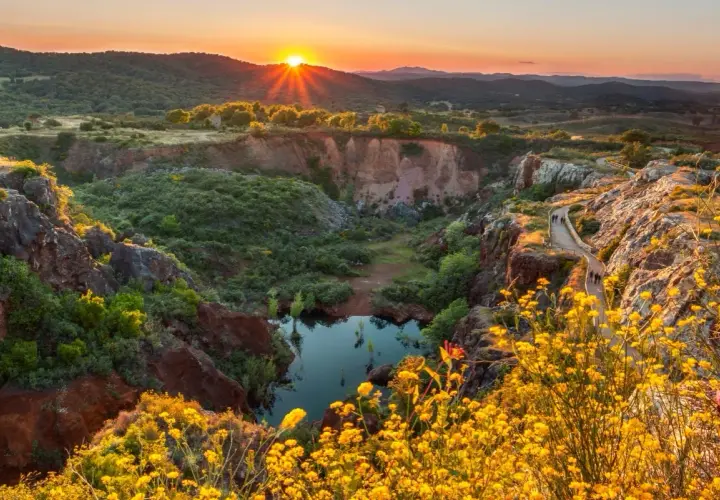 Atardecer dorado sobre un cañón con un estanque, ideal para senderismo Sevilla. Flores amarillas bordean el camino.