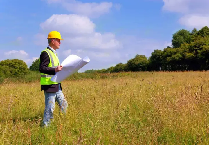 Inżynier w kasku i kamizelce odblaskowej analizuje plany na tle łąki, przygotowując się do badań geotechnicznych gruntu.
