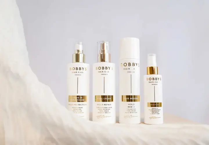 Fyra vita flaskor med guldfärgade detaljer från Bobby's Hair Care. En huskur mot statiskt hår.