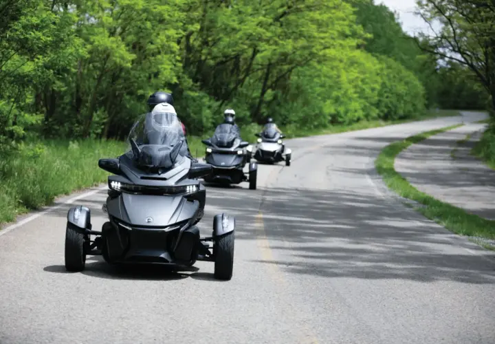 Trzy pojazdy typu Can-Am Spyder jadące po krętej drodze wśród zieleni. Ich nowoczesny design i dynamiczna jazda tworzą wrażenie efektu shimmy.