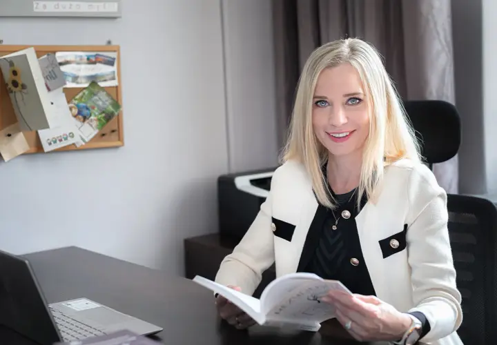 Uśmiechnięta blondynka w białej marynarce siedzi przy biurku i czyta książkę.