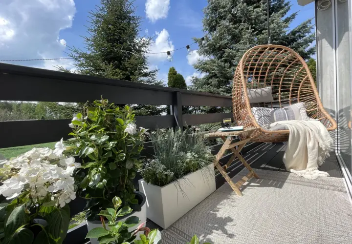 Przytulny mini balkon w bloku z fotelem wiszącym, stolikiem i kwitnącymi roślinami. Idealne miejsce na relaks.