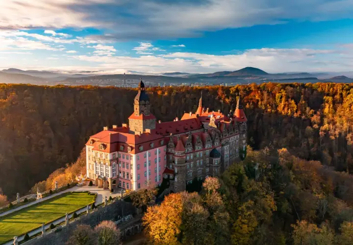 Malowniczy hotel zamek otoczony jesiennym lasem, z wieżą i czerwonymi dachami, na tle gór i błękitnego nieba.