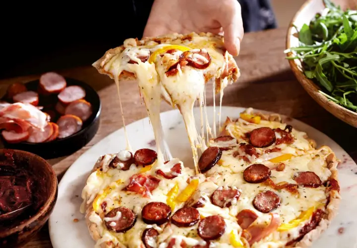 Rozciągający się ser z kawałka farmerskiej pizzy z kiełbasą i papryką. Obok miseczka z rukolą i wędlinami.