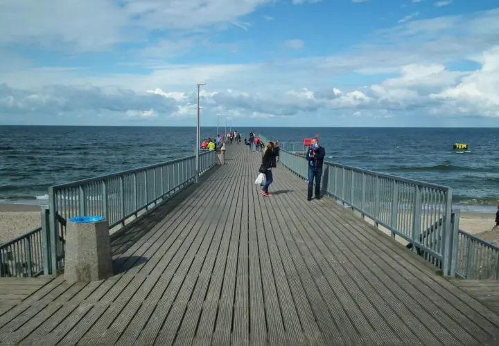 Ludzie spacerują po drewnianym deptaku nad morzem. Słońce przebija się przez chmury, a fale delikatnie uderzają o brzeg.