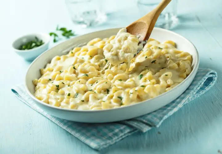 Kremowe mac n cheese z ziołami, które rozpływa się w ustach. Drewniana łyżka podnosi porcję, ukazując ciągnący się ser.