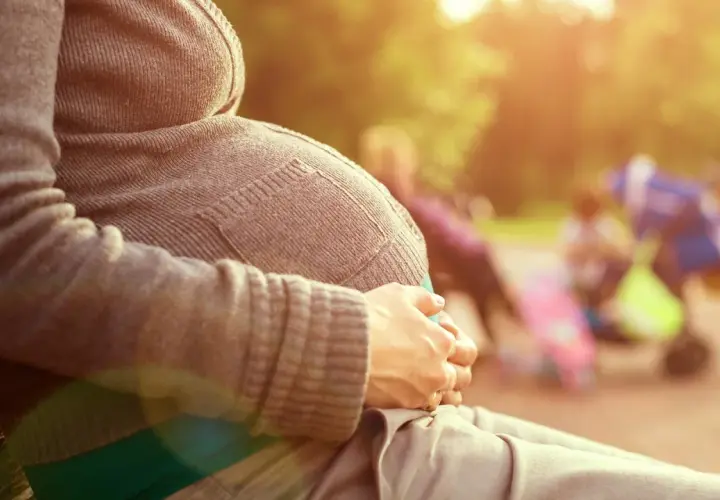 Gravid kvinna sitter på en parkbänk, njuter av solen. Hon håller sin mage, som om hon väntar på sitt barn. I bakgrunden syns andra människor och barnvagnar. Det är en varm och solig dag, med gula flytningar i luften.
