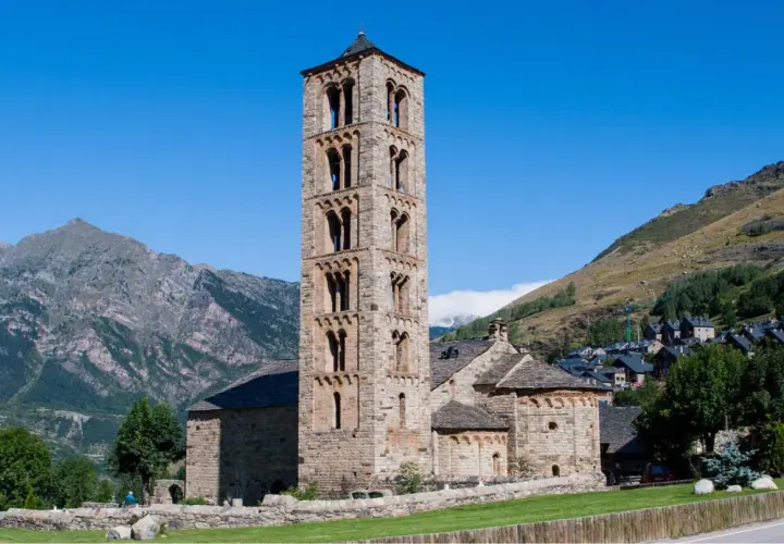 Torre románica de piedra en el Vall de Boí, con montañas y un pueblo al fondo.