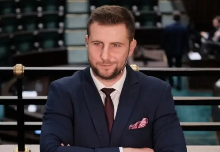 Mężczyzna w garniturze z krawatem i poszetką, siedzi na tle sali sejmowej.