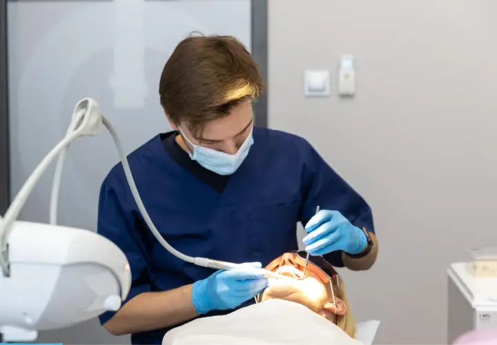 Dentysta w niebieskiej masce i rękawiczkach wykonuje zabieg. Pacjentka ma założone okulary ochronne. Nowoczesne znieczulenie komputerowe zapewnia komfort.