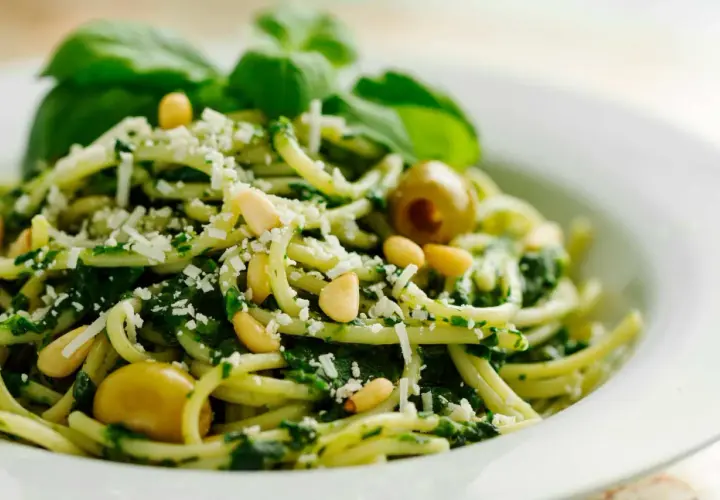 Pasta con pesto de pistacho, piñones, aceitunas verdes y queso rallado, decorada con hojas de albahaca fresca.