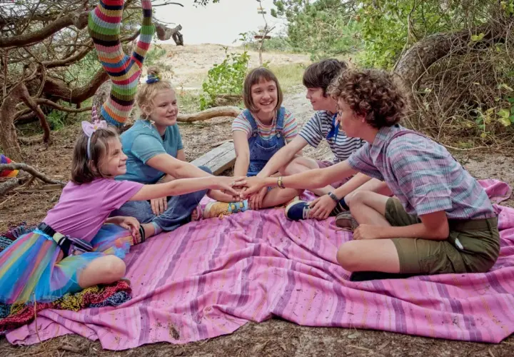 Fünf Kinder legen ihre Hände in der Mitte zusammen, bereit für ein Abenteuer. Die bunte Strickkunst am Baum deutet auf die "lotta leben reihenfolge" hin, die sie gemeinsam entdecken werden.