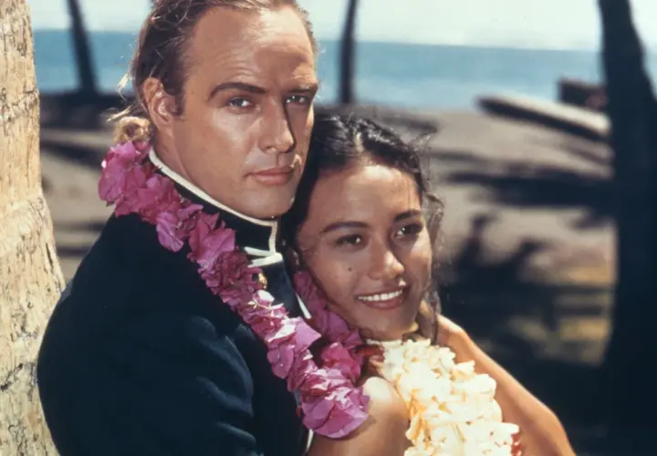 Marlon Brando z kobietą w kwiatowych girlandach na tle palmy i oceanu.