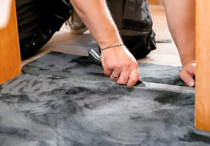 Mann schneidet mit einem Teppichmesser einen grauen Teppich zu. Das Verlegen des Teppichs wird präzise ausgeführt.