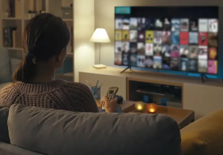 Kobieta z pilotem w ręku ogląda film na telewizorze, zastanawiając się, jak podłączyć komputer do telewizora, by cieszyć się większym ekranem.