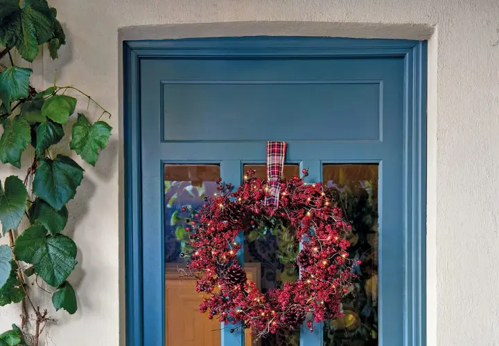 Corona navideña roja con luces y piñas colgada en una puerta azul.