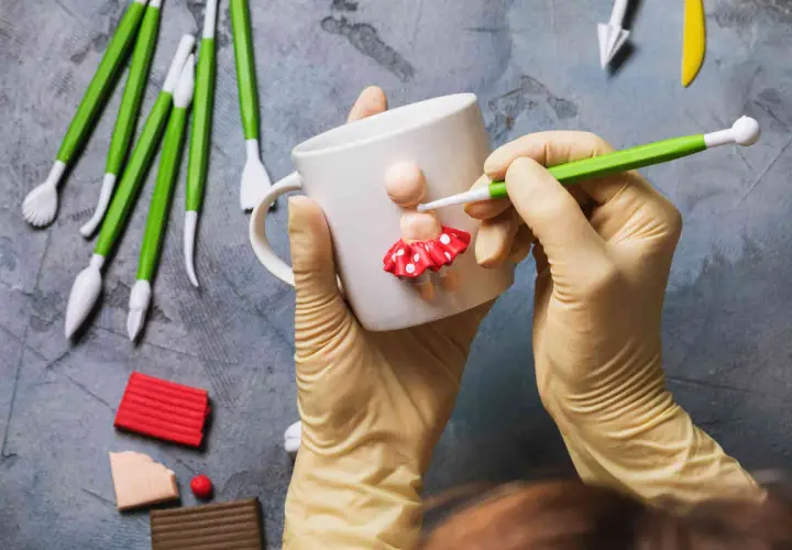 Mani guantate creano una bambolina con pasta fimo su una tazza. Tanti strumenti e colori per idee creative.