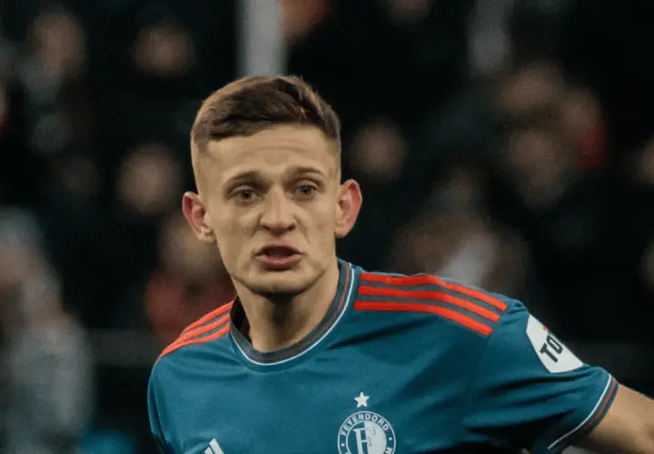 Młody piłkarz w niebieskiej koszulce z czerwonymi pasami na ramionach, z emblematem Feyenoordu. Czy to **kitman, kto to**?